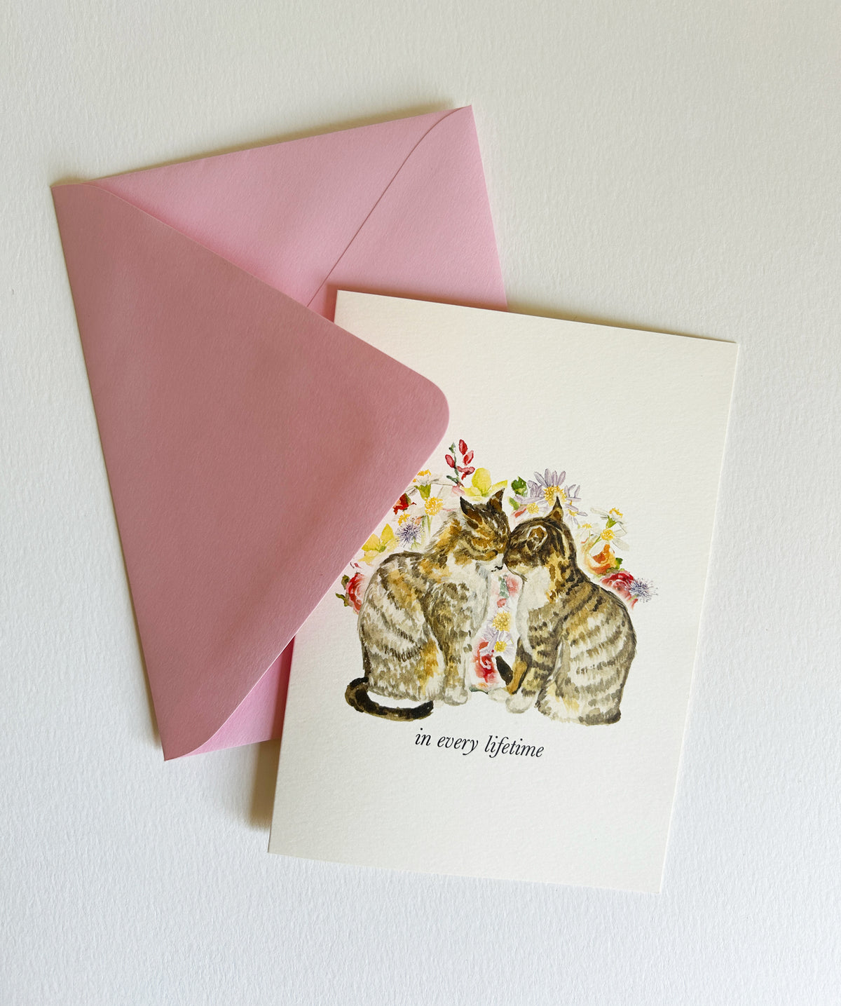 A rectangle greeting card with a pink envelope, featuring an illustration of two tabby cats surrounded by flowers with the text 'in every lifetime' on a white background.