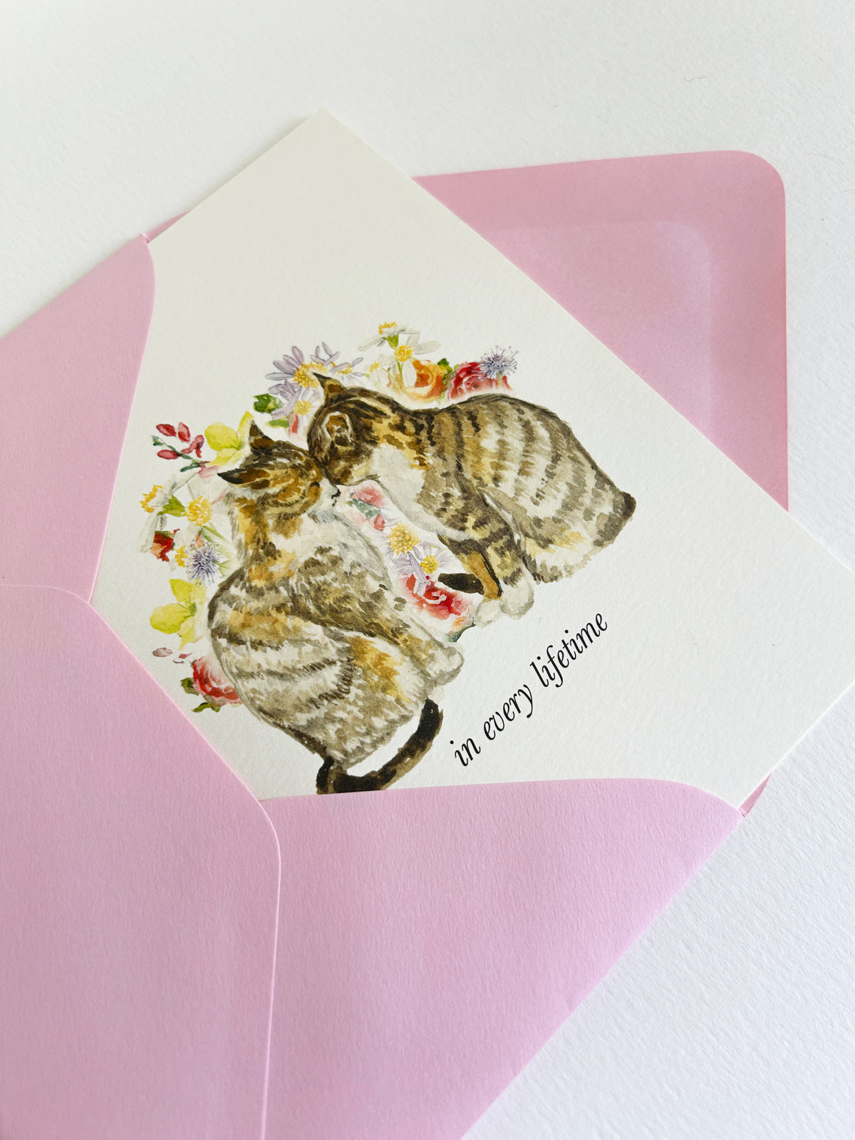 A rectangle greeting card with a watercolor illustration of two cats among small flowers, placed on top of a pink envelope. The card reads 'in every lifetime' in black serif font.