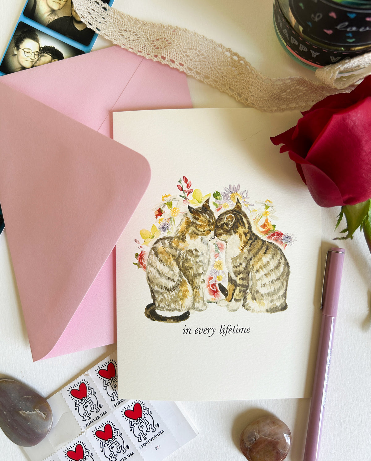 A rectangle greeting card featuring an illustration of two cats surrounded by flowers with the text 'in every lifetime', styled on a white surface with a pink envelope, a red rose, a purple pen, lace ribbon, and small heart-themed stickers.
