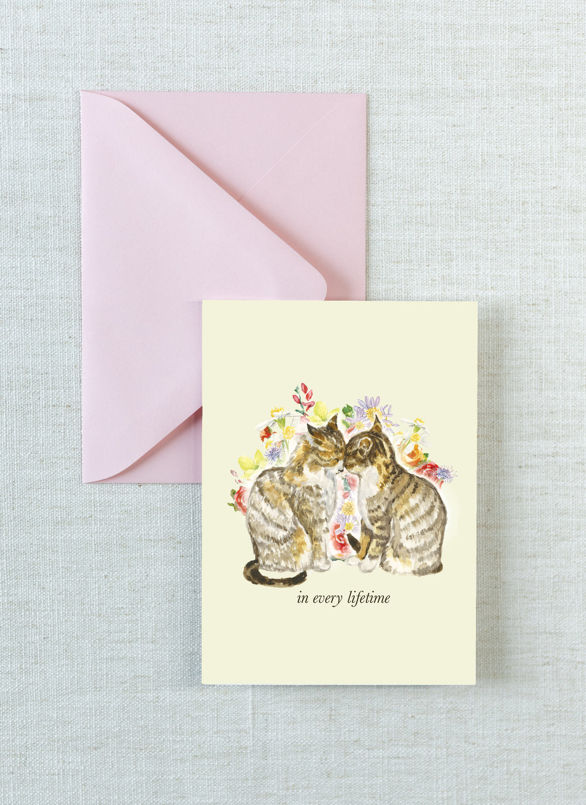 A cream-colored greeting card featuring a watercolor illustration of two cats surrounded by flowers, with the text 'in every lifetime' printed below. The card is displayed next to a light pink envelope on a textured light gray linen background.
