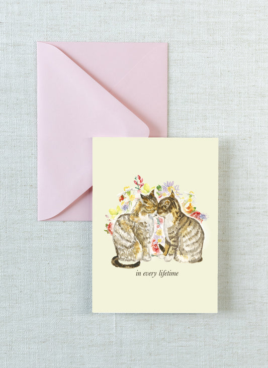 A cream-colored greeting card featuring a watercolor illustration of two cats surrounded by flowers, with the text 'in every lifetime' printed below. The card is displayed next to a light pink envelope on a textured light gray linen background.